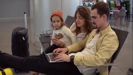 Young-father-working-on-laptop-on-airport-with-his-family-while-waiting-for-departure.-Long-haired-mother-entertains-her-little