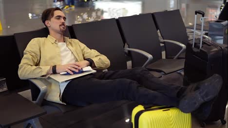 A-bearded-man-in-yellow-shirt-sleeping-holding-legs-on-the-yellow-suitcase,-holding-his-stuff-in-the-airport-lounge-while