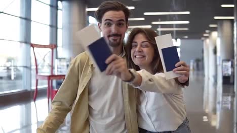 Portrait-of-excited-caucasian-couple-going-on-a-vacation.-Walking-by-airport-and-holding-passports,-boarding-passes.-Waving-with