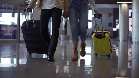 Cropped-front-footage-of-couple-travelers-are-carrying-their-luggage-and-holding-hands.-Walking-over-lounge-of-terminal.-People-are-preparing-to-boarding-and-departing-with-rolling-suitcases.-Slow-motion