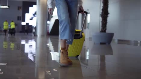 Stylish-woman-walk-with-trolley-yellow-case-by-empty-airport,-low-half-view-of-slender-female-legs-and-medium-wheeled-bag