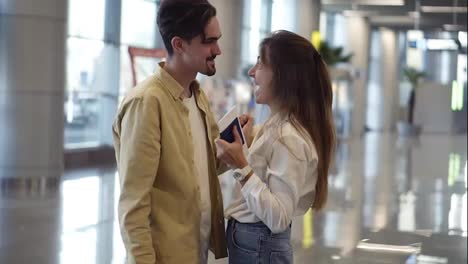 Excited,-woman-showing-passport-with-visa-stamp-to-her-boyfriend-at-the-airport.-Happy-couple,-man-hugging-his-girlfriend-and
