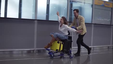 Funny-couple-in-airport.-Attractive-young-woman-and-handsome-man-with-suitcases-are-ready-for-traveling.-Having-fun-on-luggage