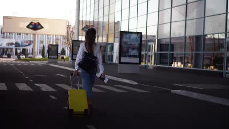 Stylish-woman-walk-with-trolley-yellow-case-by-empty-airport-terminal-outside-road,-slender-female-wearing-jeans-and-brown-boots