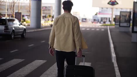 Confident,-tall-man-in-casual-with-the-black-baggage-walking-to-the-outside-airport-terminal-by-road.-Slow-motion.-Rare-view