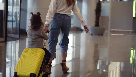 Airport-terminal,-young-unrecognizable-mother-riding-her-cute-daughter-on-a-small-yellow-suitcase.-Mom-with-yellow-suitcase-and-daughter-are-having-fun-before-their-departure