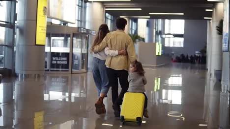 Back-view-of-modern-caucasian-family---mother,-father,-daughter---walking-the-airport-hall-and-riding-their-daughter-on-suitcase-before-the-departure-to-the-vacation.-Slow-motion