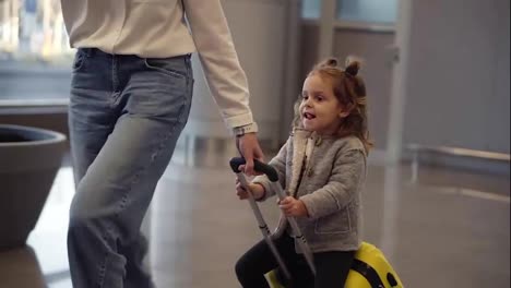 Airport-terminal,-young-unrecognizable-mother-riding-her-cute-daughter-on-a-small-yellow-suitcase.-Mom-with-yellow-suitcase-and-daughter-are-having-fun-before-their-departure.-Close-up