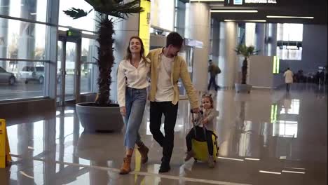 Portrait-of-smilling,-modern-caucasian-family---mother,-father,-daughter---walking-the-airport-hall-and-riding-their-daughter-on-suitcase-before-the-departure-to-the-vacation.-Slow-motion