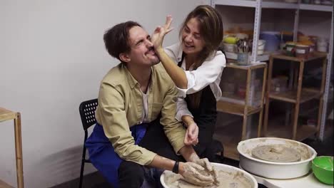 Cheerful,-smilling-couple-having-fun-while-working-together-in-potter-workshop.-Sitting-by-potter's-wheel,-woman-trying-to-clean-her-boyfriend's-face,-laughing,-excited.-Workshop-studio