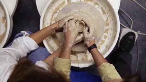 Footage-from-above-of-dirty-hands-of-romantic-couple-in-love-working-together-on-potter-wheel-and-sculpting-clay-pot,-unrecognisable-woman-and-man-mold-a-vase-in-craft-studio-workshop.-Slow-motion