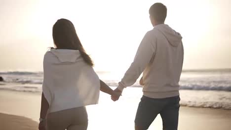 Backside-view-of-young-couple-in-love-holding-hands-walking-on-the-beach-in-casualclothes.-Background-sunset-in-sea.-Slow-Motion.-Mild-dusk