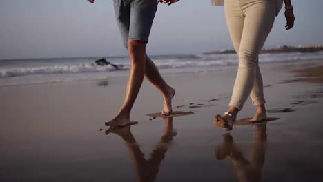 Unrecognizable-young-couple-stepping-together-at-the-golden-sand-at-sea-beach.-Male-and-female-legs-walking-near-ocean.-Bare-feet-of-pair-going-on-sandy-shore-with-waves.-Summer-vacation