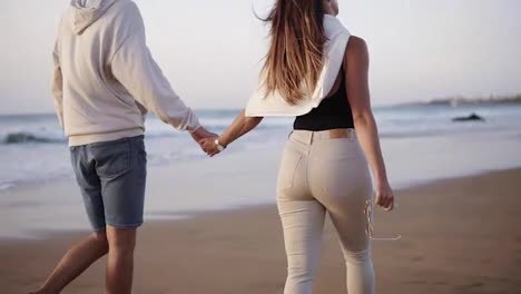 Married-young-couple-on-beach-walking-in-love-holding-hands-at-romantic-sunrise.-Woman-and-man-relaxing-on-travel-vacation-holidays,-wearing-casual-clothes.-Slow-motion.-Rare-view
