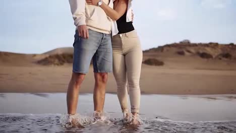 Stylish,-young-couple-standing-on-sandy-beach-shoreline-watch-sunset-waves-from-the-ocean.-Wearing-sunglsses-and-casual-clothes.-Front-view