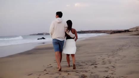Relaxed-young-couple-on-beach-walking-in-love-man-holding-his-girl-by-from-back-.-Woman-and-man-relaxing-on-travel-vacation-holidays,-man-wearing-casual-clothes,-woman-white-towel-on-belly.-Slow-motion.-Rare-view