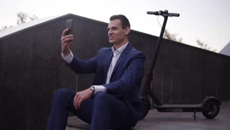 Attractive-businessman-sitting-on-the-stairs-and-does-video-chatting-on-smartphone-outside.-Man-in-elegant-blue-suit-having