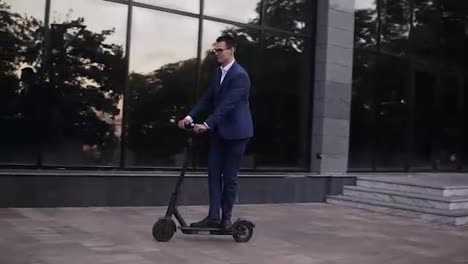 Young-businessman-in-elegant-blue-suit-and-glasses-riding-and-electric-scooter-for-a-business-meeting-in-the-office,-office