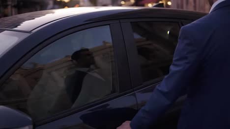 Slow-motion-young-man-walk-sitting-in-the-black-car-on-street.-Smiling-businessman-in-elegant-blue-suit-opens-the-door-of-his