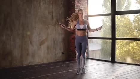 Full-length-of-a-young-blonde-athlete-woman-in-comfortable-grey-sport-outfit-jumping-rope-in-front-the-camera.-Girl-doing-jump