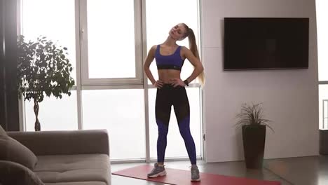Beautiful-slim-woman-in-sportswear-does-exercises-to-warm-up-and-stretch-her-neck-in-modern-airy-room-with-minimalistic-interior,-stading-close-to-blurred-panoramic-windows