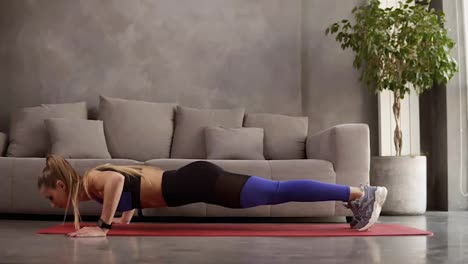 Healthy-young-woman-in-sportswear-doing-push-ups-on-fitness-mat-in-front-the-sofa.-Young-female-exercising-at-home.-Side-view
