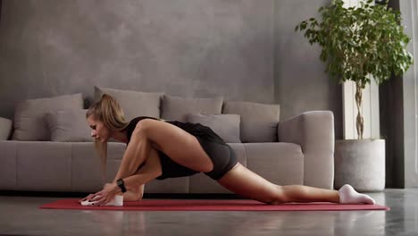Longhaired,-blonde-woman-in-black-sportswear-trainings-at-home-on-mat-in-front-the-grey-sofa-and-stretching-muscles-of-legs,-making-lunge,-leaning-torso-forward.-Loft-interior-room