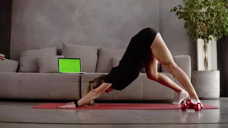 Caucasian,-blonde-woman-in-black-sportswear-trainings-at-home-on-mat-in-front-the-grey-sofa-and-stretching-muscles-of-legs,-sitting-on-floor,-leaning-torso-forward.-Loft-interior-room.-Dumbbells-on-the-floor-close-to-the-mat