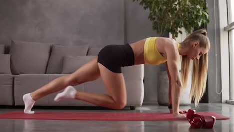 Young-fit-sporty-blonde-girl-with-pigtail-in-yellow-top,-black-shorts-does-crossing-legs-exercises-in-plank-position-on-mat-in-bright-room.-Doing-sports-and-fitness-training-at-home.-Healthy-lifestyle,-home-fitness