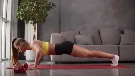 Healthy-young-woman-in-sportswear---yellow-top-and-black-shorts-doing-push-ups-on-fitness-mat-in-front-the-grey-sofa-in-living-room.-Young-female-exercising-at-home.-Side-view