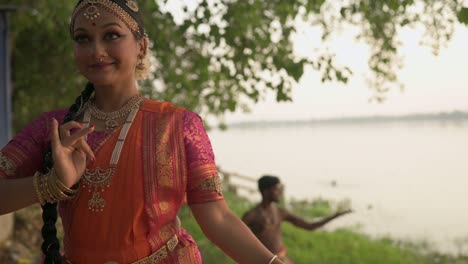 Two-Indian-classical-dancers-dancing-bharatnatyam,-Beautiful-indian-woman-and-man-dancing-at-ganga-ghat-or-riverbank,-slow-motion
