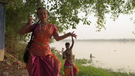 Two-dancers-dancing-Indian-classical-dance-bharatnatyam-at-Ganga-river-bank-at-twilight,-slow-motion