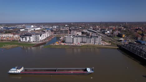 Large-empty-cargo-ship-in-the-foreground-passing-by-on-river-IJssel-in-front-of-Noorderhaven-neighbourhood-in-Zutphen,-The-Netherlands,-seen-from-above