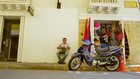 Man-sits-reads-paper-by-blue-motorbike-outside-shop-in-Cartagena