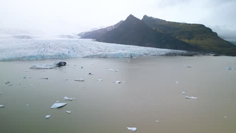 Drohne-Fliegt-über-Eisberg-Mit-Gletscher-Und-Grünem-Berg-Im-Hintergrund