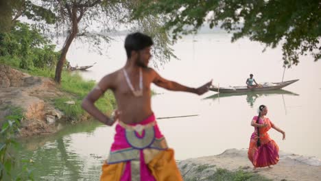 Indische-Tänzerinnen-Und-Tänzer-Tanzen-Den-Klassischen-Indischen-Tanz-Bharatnatyam-Am-Flussufer,-Boot-Im-Hintergrund,-Zeitlupe