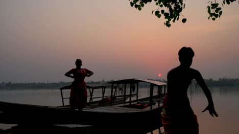 Eine-Silhouette-Einer-Männlichen-Und-Weiblichen-Tänzerin,-Die-Den-Klassischen-Indischen-Tanz-Bharatnatyam-Auf-Einem-Boot-Bei-Sonnenuntergang-Oder-Sonnenaufgang-Am-Ganga-Flussufer-Tanzt,-Zeitlupe