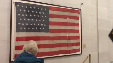 Mirando-Una-Vieja-Bandera-Estadounidense-Enmarcada-Colgada-En-La-Pared-Del-Edificio-De-La-Capital-Del-Estado-De-Utah,-Y-Una-Mujer-Rubia-Pasa-Caminando