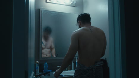 Man-walks-out-towards-mirror,-towel-wrapped-around,-wipes-steam-off-and-views-his-reflection-in-the-mirror