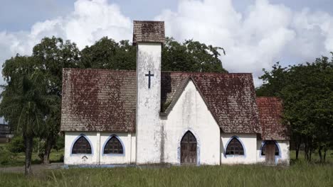 An-old-abandoned-white-and-blue-Christian-chapel-with-a-small-cross-boarded-up-surrounded-by-weeds-and-tropical-trees-on-the-side-of-a-highway-in-the-state-of-Pernambuco-in-Northeastern-Brazil