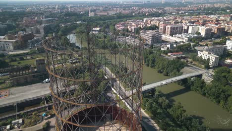 Aerial-drone-shot-flying-high-over-an-old-iron-structure,-Gazometro-in-Ostiense-district,-Rome,-Italy-with-the-view-of-the-city-in-the-background