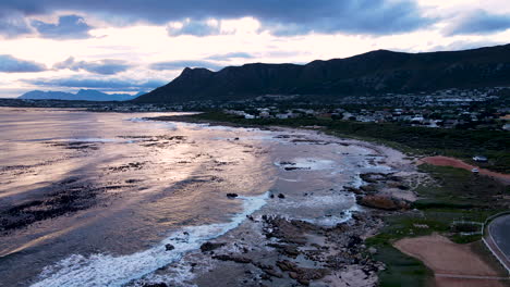 Sunset-drone-view-of-Sandbaai-coastline-with-swell-rolling-in,-mountain-backdrop