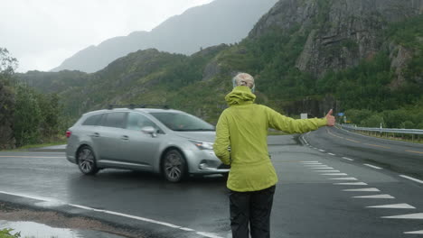 Pretty-Blonde-Girl-Hitchhiking-at-the-Side-of-the-Road-in-the-Lofoten-Islands-on-a-Cold-Cloudy-Rainy-Day,-Cars-Are-Passing,-Norway