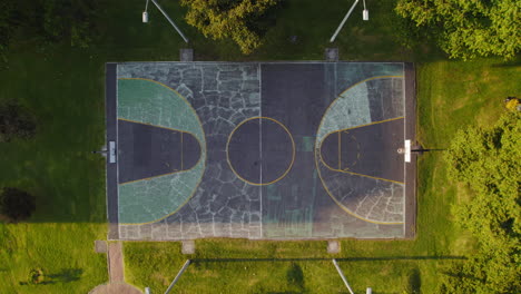 Aerial-Drone-View-Of-Cracked-Concrete-Empty-Basketball-Court-In-Outdoor-Park-Surrounded-By-Lush-Green-Grass-And-Trees-During-Sunny-Sunrise-Sunset
