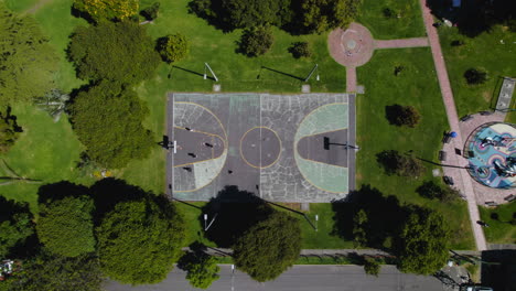 Draufsicht-Auf-Menschen,-Die-Basketball-Auf-Einem-Betonierten-Basketballplatz-Im-Außenpark-Mit-üppigem-Grünem-Gras-Und-Bäumen-Spielen