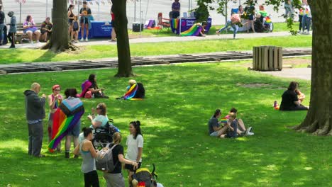 Aerial-view-showing-resting-people-on-grass-area-in-park-with-LGBT,-Gay-Pride-And-Lesbian-Pride-Flags