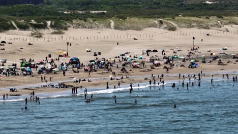Crowds-of-People-Enjoy-Summer-Vacation-on-Scheveningen-Beach,-Swimming-in-the-Sea-and-Tanning-Lying-on-Sand,-Holland---Aerial-Parallax-Movement