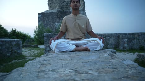 Junger-Indischer-Mann-öffnet-Seine-Chakren-Mit-Tiefer-Meditation-Und-Hatha-Yoga,-Während-Er-Bei-Sonnenaufgang-Auf-Einer-Steinmauer-Einer-Burgruine-Auf-Dem-Hügel-Sitzt