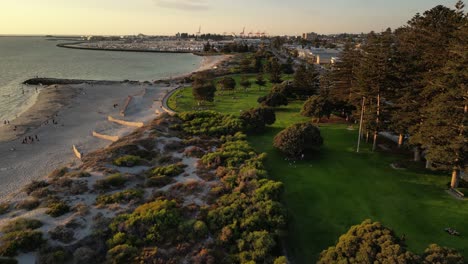 Aerial-establishing-drone-flight-over-dunes,-sandy-beach-and-South-Beach-Park-in-Fremantle,-Western-Australia---Harbor-Port-in-background-during-golden-sunset