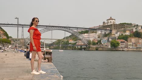 Mujer-Turista-En-El-Paseo-Marítimo-De-Ribeira-Con-El-Puente-Dom-Luís-I-Al-Fondo-En-Porto,-Portugal